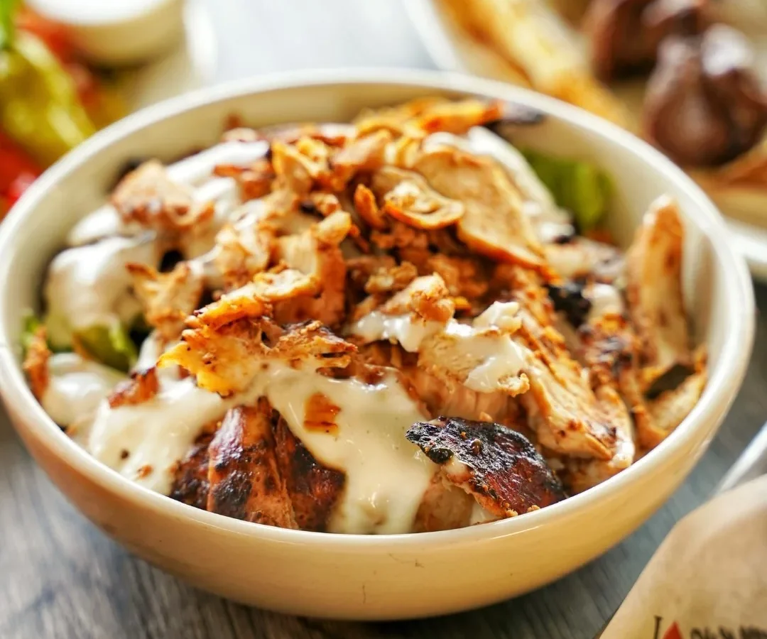 A bowl of shaved chicken shawarma over greens with garlic sauce and crispy onions, with kabob skewers and fries alongside