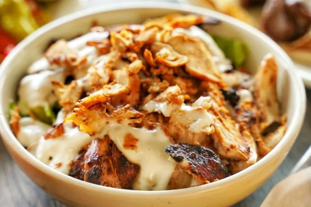 A bowl of shaved chicken shawarma over greens with garlic sauce and crispy onions, with kabob skewers and fries alongside
