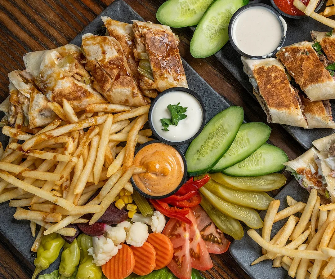 Two slate boards of sliced Abu Omar Halal shawarma with seasoned fries, pickles, cucumber, tomato, olives and dipping sauces