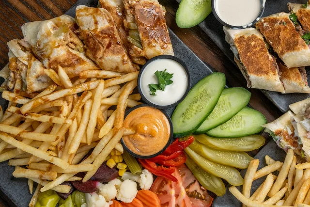Two slate boards of sliced Abu Omar Halal shawarma with seasoned fries, pickles, cucumber, tomato, olives and dipping sauces